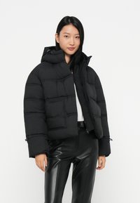 Zwarte cropped pufferjack met een capuchon, voorzien van gestikte vierkanten, een rits aan de voorkant, en trekkoorden op de mouwen, gedragen met zwarte leren broek.