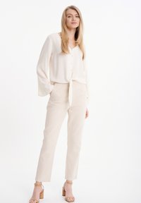 Blusa de color beige claro con lazo delantero y mangas largas, combinada con pantalones de lino ajustados a juego y sandalias de tacón cuadrado en tono marrón claro.