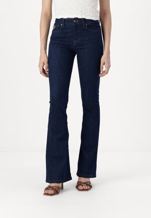 Frau trägt hoch taillierte, dunkelblaue Schlagjeans kombiniert mit braunen, mit Nieten besetzten Sandaletten mit offenem Zehenbereich, steht vor einem schlichten weißen Hintergrund.