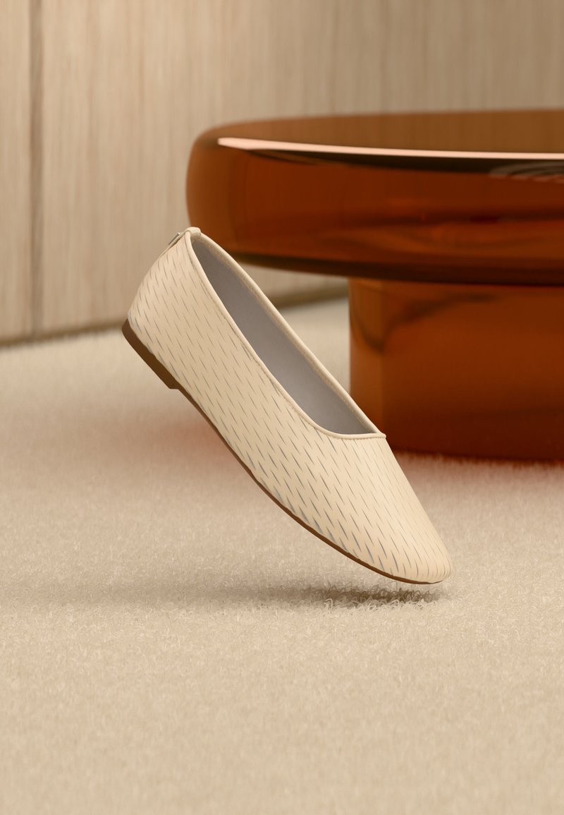 Μπεζ επίπεδη παπούτσια τύπου slip-on με διακριτικό διαγώνιο σχέδιο ραφής, τοποθετημένα πάνω σε υφασμένο χαλί, με στρογγυλό τραπέζι χρώματος κεχριμπαριού στο φόντο.