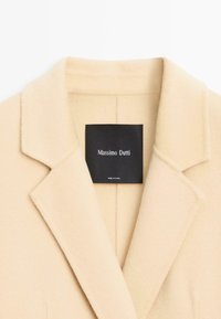 Beige blazer i uldblanding med hakket krave, enkeltknappet design og et sort mærke med "Massimo Dutti." Blød tekstur.