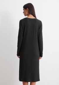 Dlouhá černá šaty s dlouhými rukávy, ribbovanou texturou, kulatým výstřihem a bočními rozparky pro volný pohyb. Jednoduchý a minimalistický design.