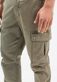 Olivgrüne Cargo-Hose aus Baumwolle mit Seitentaschen und einem sichtbaren Markenlogo-Patch. Die Textur wirkt glatt mit verstärkten Nähten.