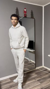 Lichtgrijze hoodie en bijpassende joggingbroek met een ontspannen pasvorm. Witte sneakers, staand naast een spiegel en een donkere tv op een plank.