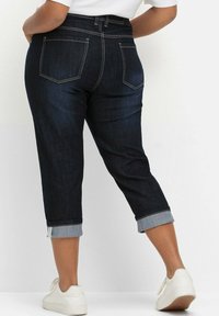 Tmavě modré džíny capri s ohrnutými nohavicemi a kontrastním stehem. Obsahuje dvě zadní kapsy a vysoký pas. Nenošeny s bílými botami.