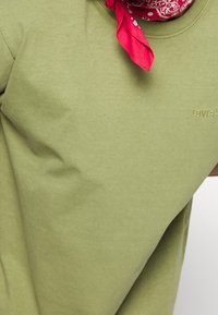 T-shirt en coton vert olive avec un col rond, présentant un petit logo brodé de Levi's. Un bandana rouge est noué autour du cou.