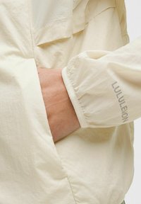 Lys beige vandtæt jakke med tekstureret overflade, elastiske manchetter og synlig logo på ærmet. Har en frontlomme.