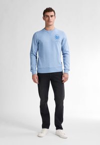 Lichtblauwe sweatshirt met ronde hals, ribgebreide manchetten en zoom; voorzien van een bedrukte graphic op de borst; gecombineerd met donkere spijkerbroek en witte sneakers.