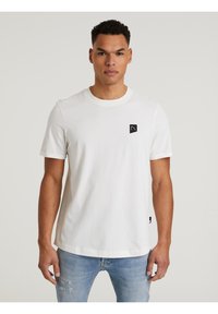 Weißes Baumwoll-T-Shirt mit kurzen Ärmeln, das ein schwarzes Logo-Patch auf der linken Brust und einen geraden Saum aufweist. Getragen mit hellblauen, ausgebleichten Jeans.