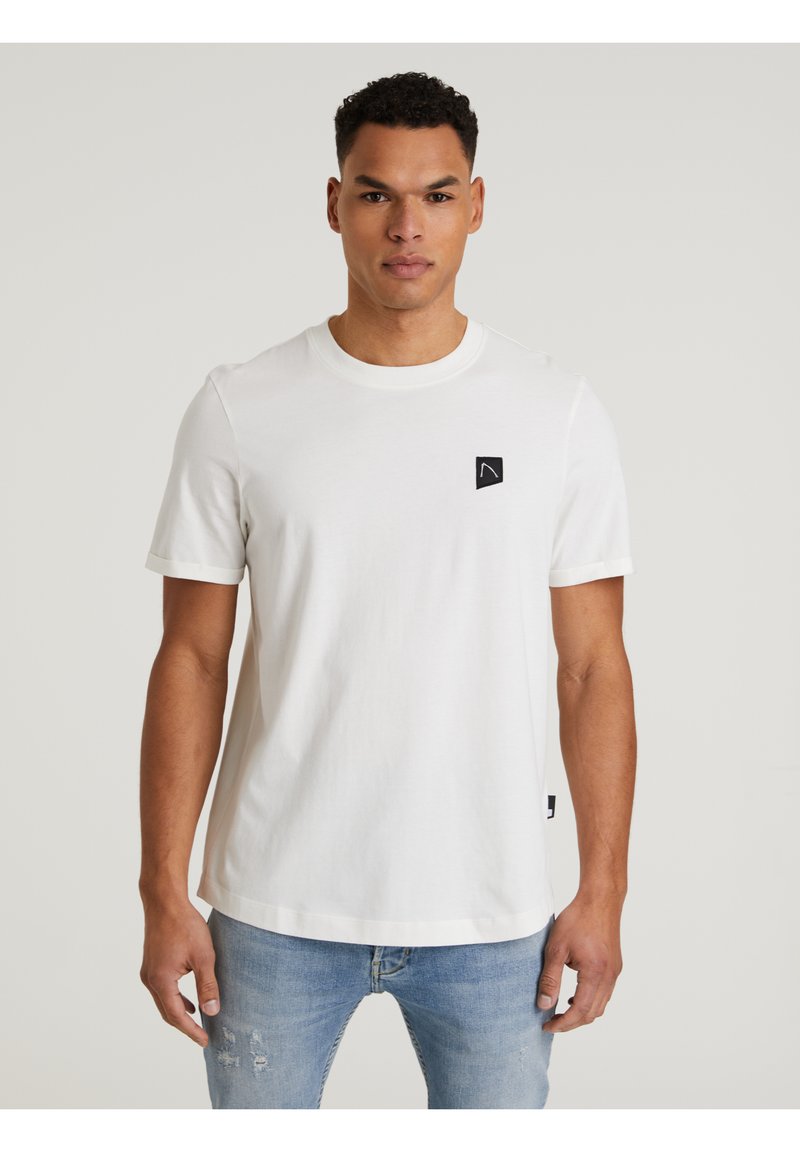 Weißes Baumwoll-T-Shirt mit kurzen Ärmeln, das ein schwarzes Logo-Patch auf der linken Brust und einen geraden Saum aufweist. Getragen mit hellblauen, ausgebleichten Jeans.