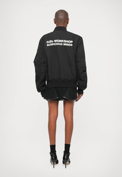 Veste bomber noire avec fermeture éclair sur le devant, col côtelé et manches surdimensionnées. Présente un texte blanc au dos et une finition lisse et mate.