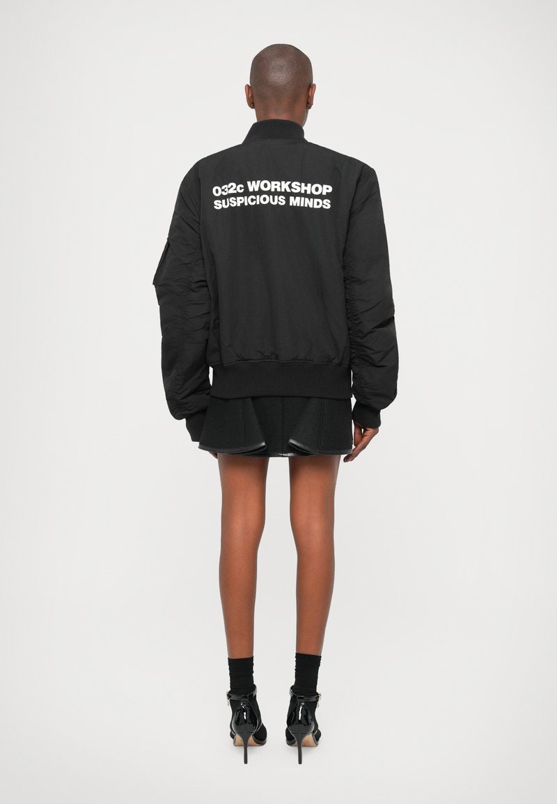 Veste bomber noire avec fermeture éclair sur le devant, col côtelé et manches surdimensionnées. Présente un texte blanc au dos et une finition lisse et mate.