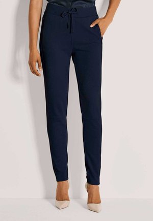 Vrouw in navy slim-fit drawstring broek en beige puntige hakken, staand tegen een effen lichte achtergrond.