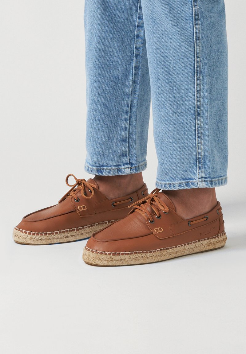 Brown leather boat shoes with lace closure, jute midsole, and textured sole; paired with light blue cuffed denim pants.