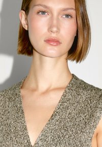 Junge Frau mit kurzem, hellbraunem Haar, die ein gestricktes ärmelloses Oberteil in neutralen Tönen trägt, vor einem einfarbigen Hintergrund.