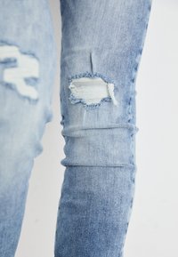 Jean délavé bleu clair avec des pièces effilochées aux genoux, offrant une coupe slim et une texture légèrement décolorée.