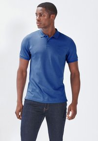 Polo azul hecho de una mezcla de algodón, con un cuello clásico, mangas cortas y un logo sutil en el pecho. Llevado con jeans oscuros.