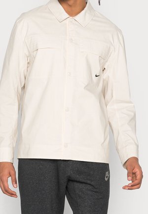 Man wearing a cream button-up jacket with chest pockets and dark grey Nike sweatpants against a plain background.