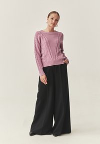 Pull en tricot torsadé rose clair avec un motif en V, associé à un pantalon noir à jambes larges et des talons noirs. Texture lisse, coupe décontractée.