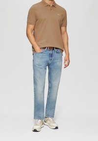 T-shirt polo marron, jean droit bleu clair et baskets beigе. La chemise présente un col et un détail de logo brodé.