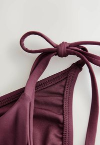 Bordowy stanik bikini z regulowanymi paskami materiałowymi związanymi w kokardkę, gładką teksturą oraz przeszywaną krawędzią.