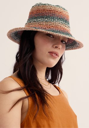 Young woman with long dark hair wearing a multicolored woven hat and orange sleeveless top, looking slightly sideways with a neutral expression.