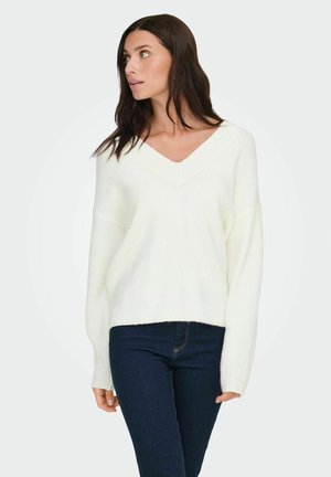 Femme en pull blanc à col en V et manches longues et jean bleu foncé regardant vers la gauche sur fond uni.
