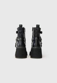 Botas de tornozelo de couro preto com saltos robustos, correias decoradas com estrelas e alças para puxar, vistas de trás sobre um fundo branco liso.