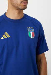 Kék sportpóló arany Adidas csíkokkal, hímzett Italia címer zöld, fehér és piros részletekkel, valamint négy arany csillaggal. Sima textúra.