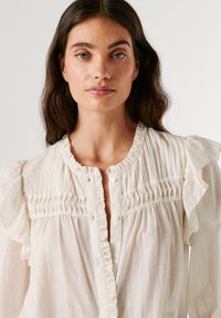 Šviesiai kreminė bluzelė, pasižyminti raukytomis rankovėmis, sagų priekyje ir vertikaliomis klostėmis per krūtinę. Lygios, tekstūruotos medžiagos. Paprastas dizainas.