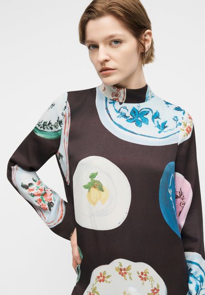 Γυναίκα με κοντά μαλλιά που φοράει μαύρο φόρεμα με ψηλό λαιμό, διακοσμημένο με μεγάλα πολύχρωμα πιάτα με σχέδια από λουλούδια και φρούτα.