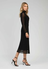 Robe noire à manches longues avec un corsage ajusté, texture en maille transparente et longueur midi. Associée à des sandales à talons hauts noires à lanières.