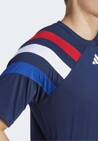 Námořnické sportovní tričko s krátkým rukávem, které má červené, bílé a modré pruhy přes ramena. Vyrobeno z hladkého, prodyšného materiálu.