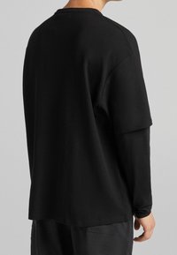 Osoba v černém texturovaném krátkém rukávu tričku, které má přes něj oblečené černé dlouhé rukávy tričko, viděná zezadu na světlém šedém pozadí.