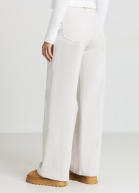 Weit geschnittene, elfenbeinfarbene Cordhosen mit einem strukturierten Muster. Verfügen über Gesäßtaschen und einen elastischen Bund. Kombiniert mit beigen Schuhen.