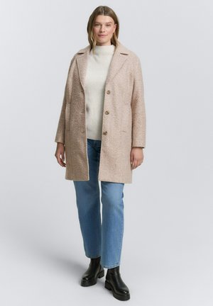 Modèle féminin portant un manteau beige texturé sur un pull blanc côtelé, un jean bleu clair et des bottes noires épaisses, debout devant un fond gris.