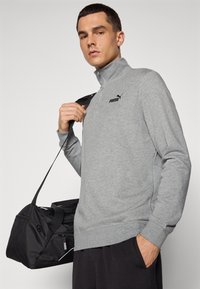 Šedý svetr s dlouhým rukávem a límcem na zip, se znakem Puma v černé barvě. Model drží černou sportovní tašku se zipy.