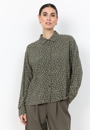 Woman wearing an olive green button-up shirt with white polka dots and matching olive pants, standing with one hand in pocket.