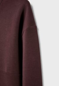 Burgundy collegepaita pehmeällä tekstuurilla, jossa on pudotettu olkapäädesign ja etutasku. Saumadetaali näkyy vasemmalla puolella.