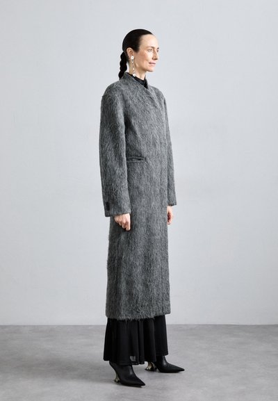 Palton lung gri din material pufos, cu buzunare laterale, purtat peste o rochie neagră în straturi și cizme negre cu bot ascuțit. Design simplu al gulerului.