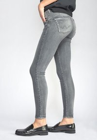 Osoba v úzkých bledě šedých džínách a černých mokasínách, postavená bokem s jednou rukou v kapse na jednobarevném pozadí.