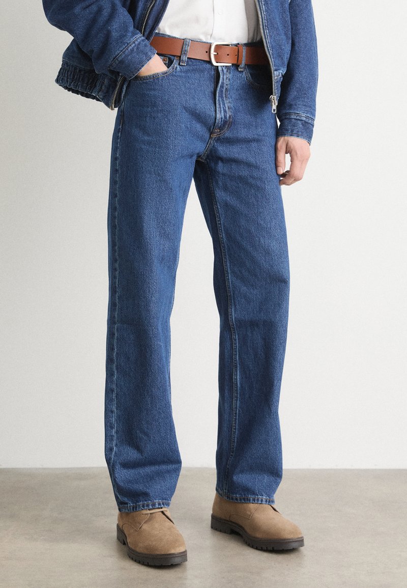 Jeans en denim bleu avec une coupe décontractée, comprenant cinq poches, des passants de ceinture et une ceinture en cuir brun visible, associés à des bottes en daim marron.