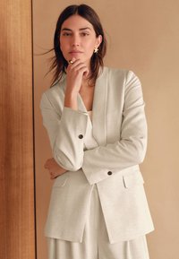 Lichtgrijs colbert met sjaalkraag, zijzakken en manchetten met knopen, gecombineerd met bijpassende broek. Soepele, gestructureerde stof.