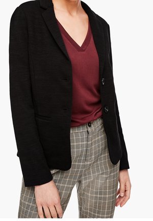 Femme portant un blazer noir, un haut bordeaux à col en V et un pantalon à carreaux beige, la main détendue le long du corps, sur un fond blanc.