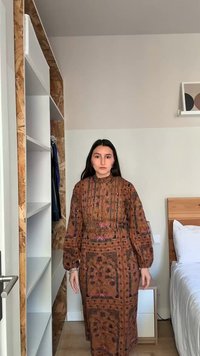 Kvinde med langt mørkt hår iført en brun mønstret langærmet kjole, der står i et minimalistisk soveværelse med hvide vægge og møbler i træ.