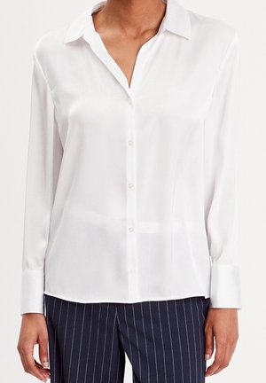 Chemise blanche à manches longues avec col boutonné portée par une personne, associée à un pantalon bleu marine foncé à fines rayures.