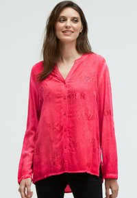 Frau in leuchtend pinkfarbener, langärmliger Bluse mit Knöpfen und dezent bedrucktem Text, kombiniert mit schwarzen Hosen, die vor einem hellen Hintergrund steht.