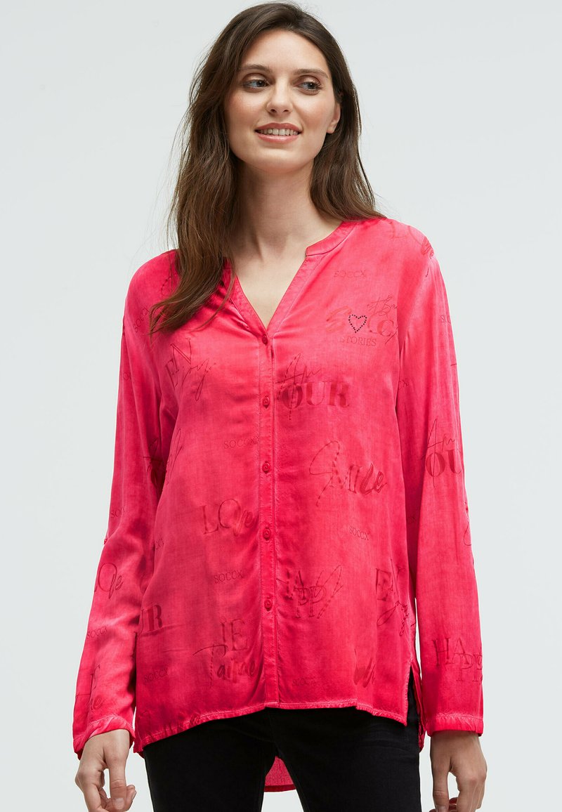 Frau in leuchtend pinkfarbener, langärmliger Bluse mit Knöpfen und dezent bedrucktem Text, kombiniert mit schwarzen Hosen, die vor einem hellen Hintergrund steht.