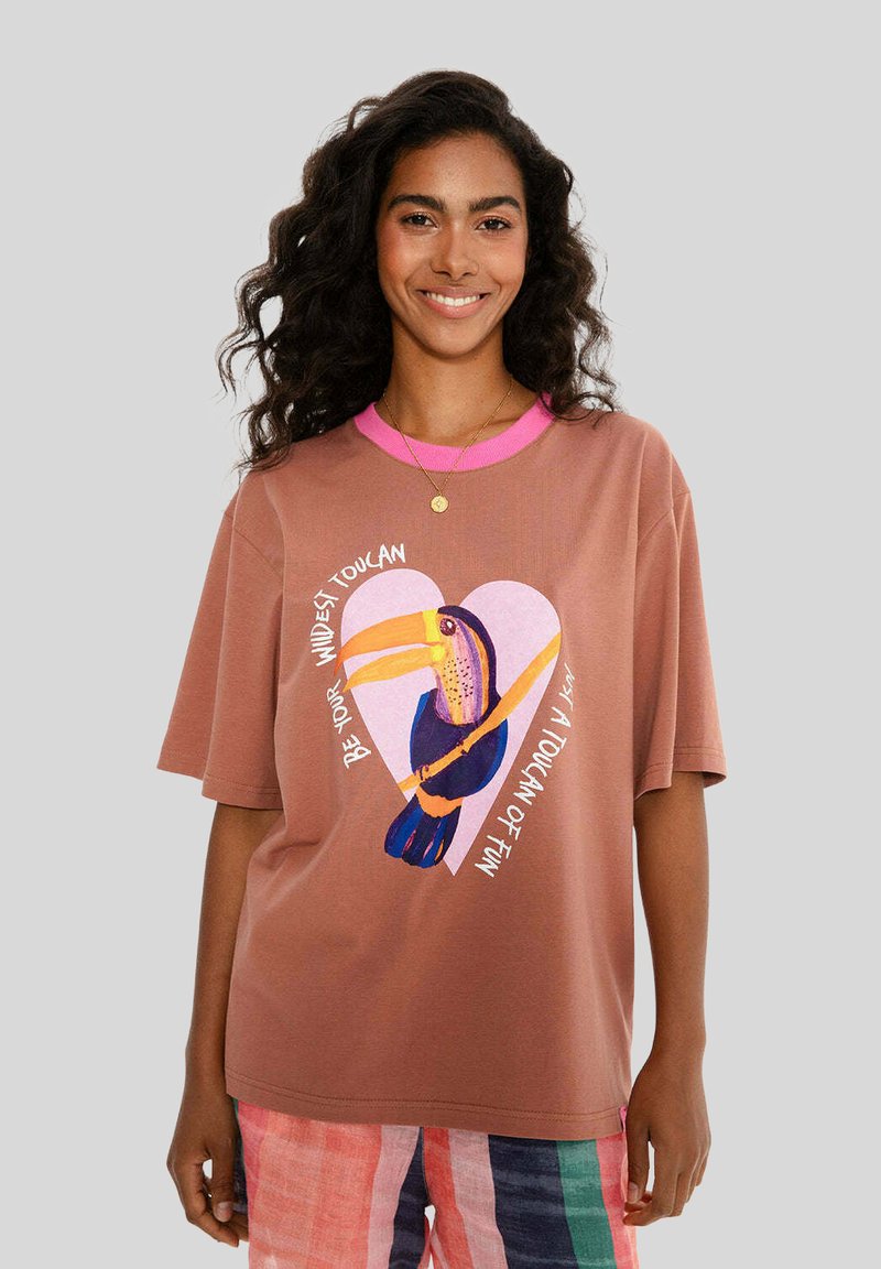 Hnědý tričko s krátkým rukávem a růžovým výstřihem, s barevnou grafikou tukana uvnitř srdce a textem kolem něj. Měkká bavlněná tkanina.