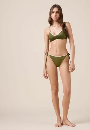 Femme debout pieds nus portant un bikini vert olive avec une bordure blanche, noué aux hanches, sur un fond beige uni.